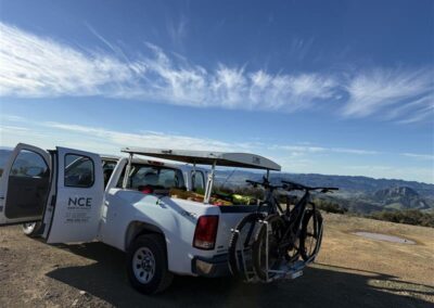 A white truck with doors open is parked upon a dirt clearing overlooking mountains, clouds, and a blue sky.