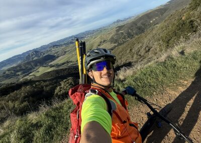 A man with orange safety vest, neon green shirt, sunglasses, and a bike helmet takes a selfie atop his mountain bike with survey equipment in his backpack. Dirt is on the ground and rolling green hills are in the background.