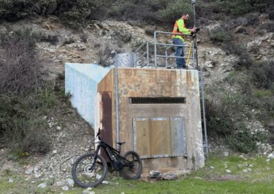 Man in neon green shirt and orange safety vest sets up survey equipment atop a concrete structure with a mountain bike leaned against it.