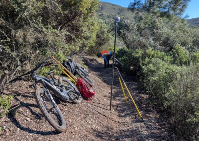 Mountain bikes and survey equipment on a trail surrounded by green bushes.